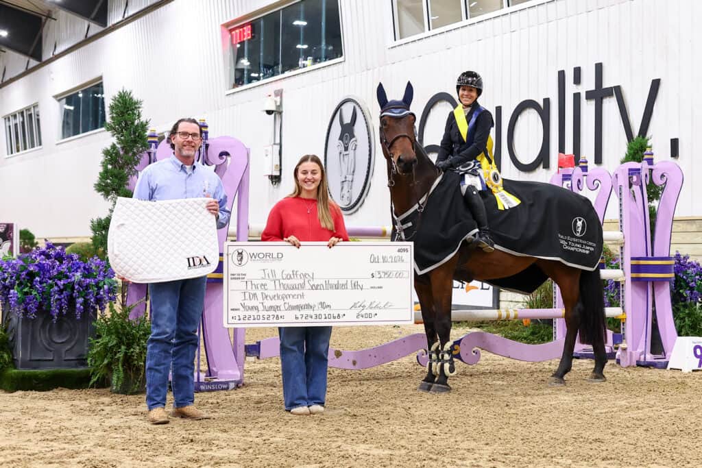 A man, a woman, and a rider on a horse stand together in an equestrian arena holding a large check for $3,750.00 awarded to Jill Gaffney at the World Equestrian Center.