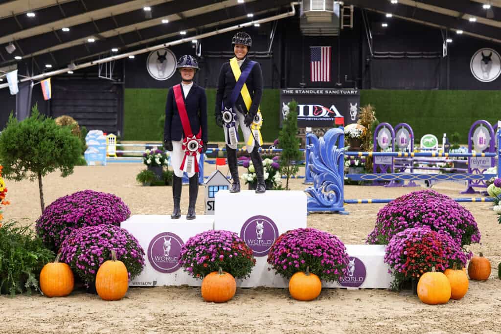 Two equestrians in riding gear stand on a podium with ribbons, surrounded by mums and pumpkins in an indoor arena.