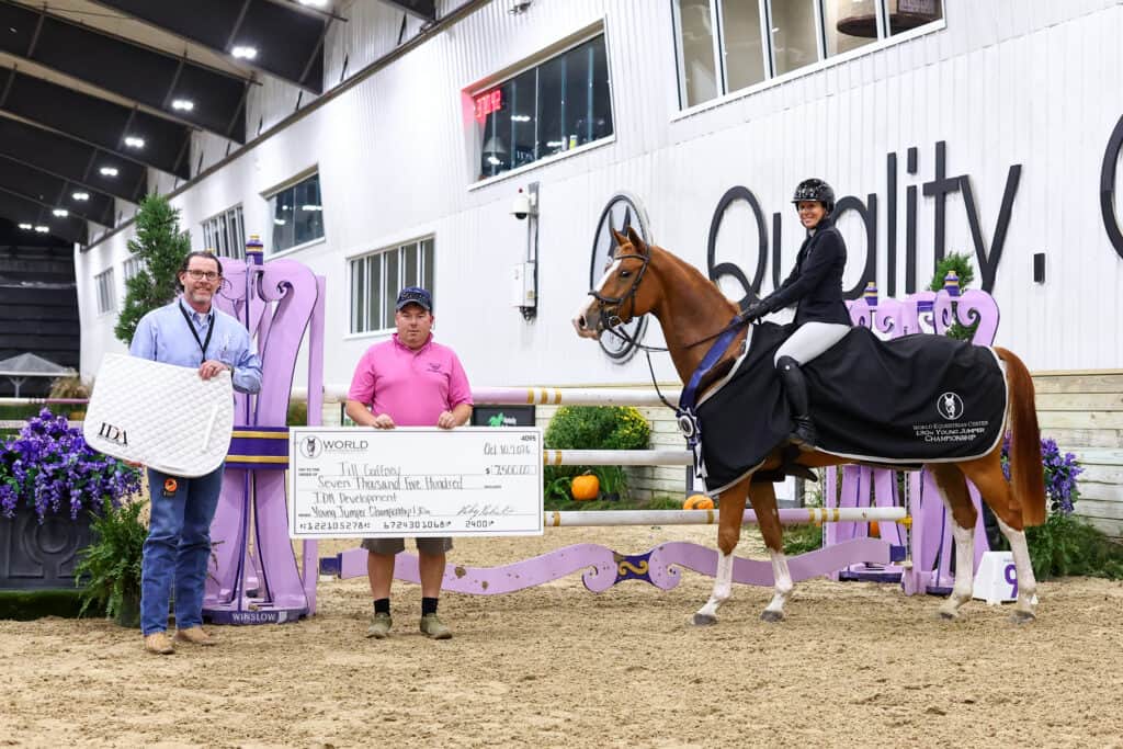 A man in a blue shirt, a man in a pink polo holding a large check, and a woman on a horse wearing a winner’s blanket in an indoor equestrian arena.