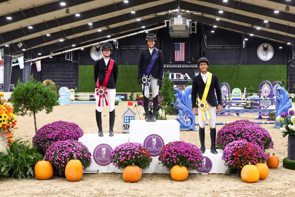 Three equestrian riders in formal show attire standing on a winner’s podium decorated with purple mums and pumpkins.