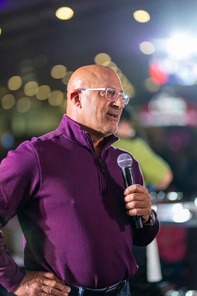 A man in a purple quarter-zip sweater speaks into a handheld microphone.
