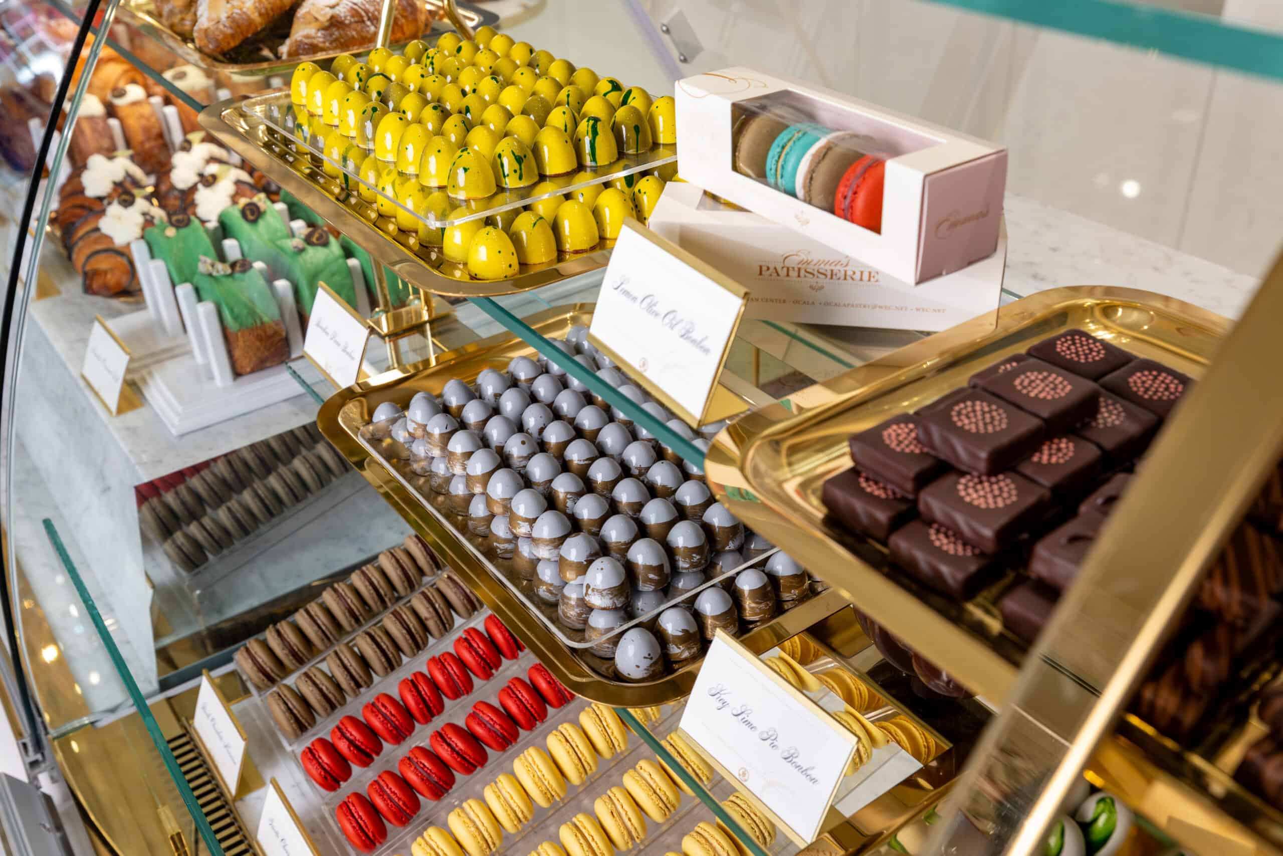 Glass display case filled with rows of colorful chocolates, macarons, and pastries on golden trays.