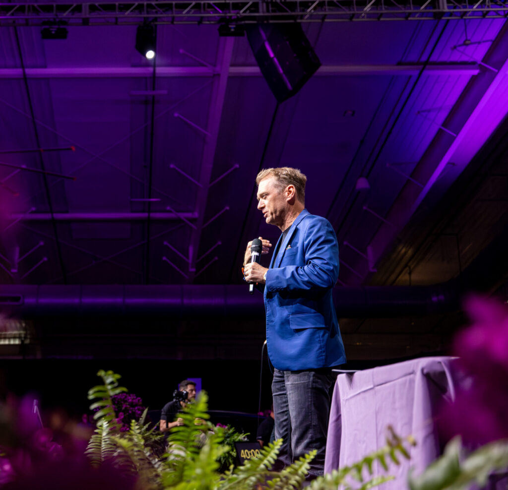 A man in a blue blazer speaks into a microphone on a stage lit with purple light.