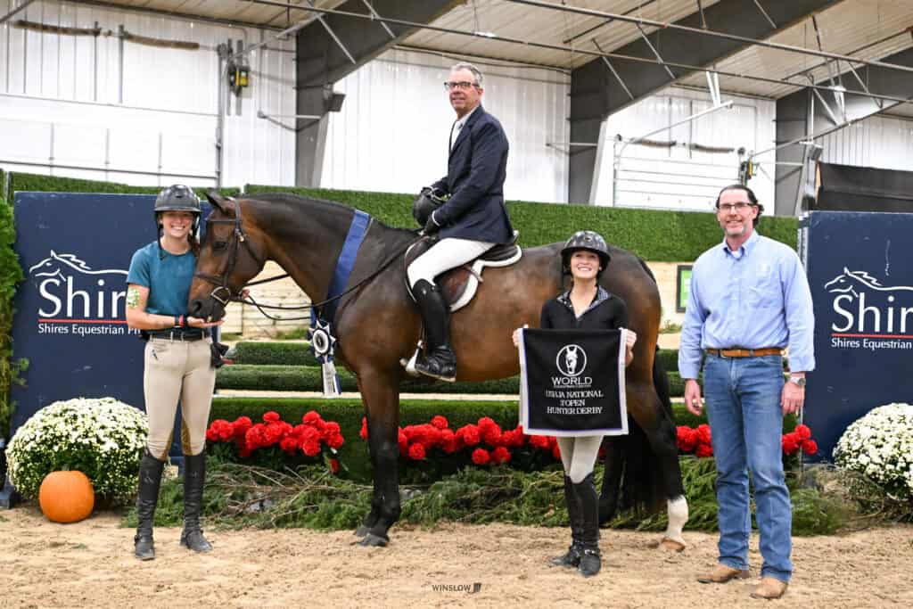 A rider on a brown horse stands between two people in an indoor arena, with one person holding a World Championship Hunter Derby winner’s banner.