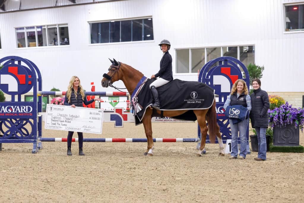 A rider on horseback stands in an arena with three women holding a check for $18,000 and a sponsor blanket.