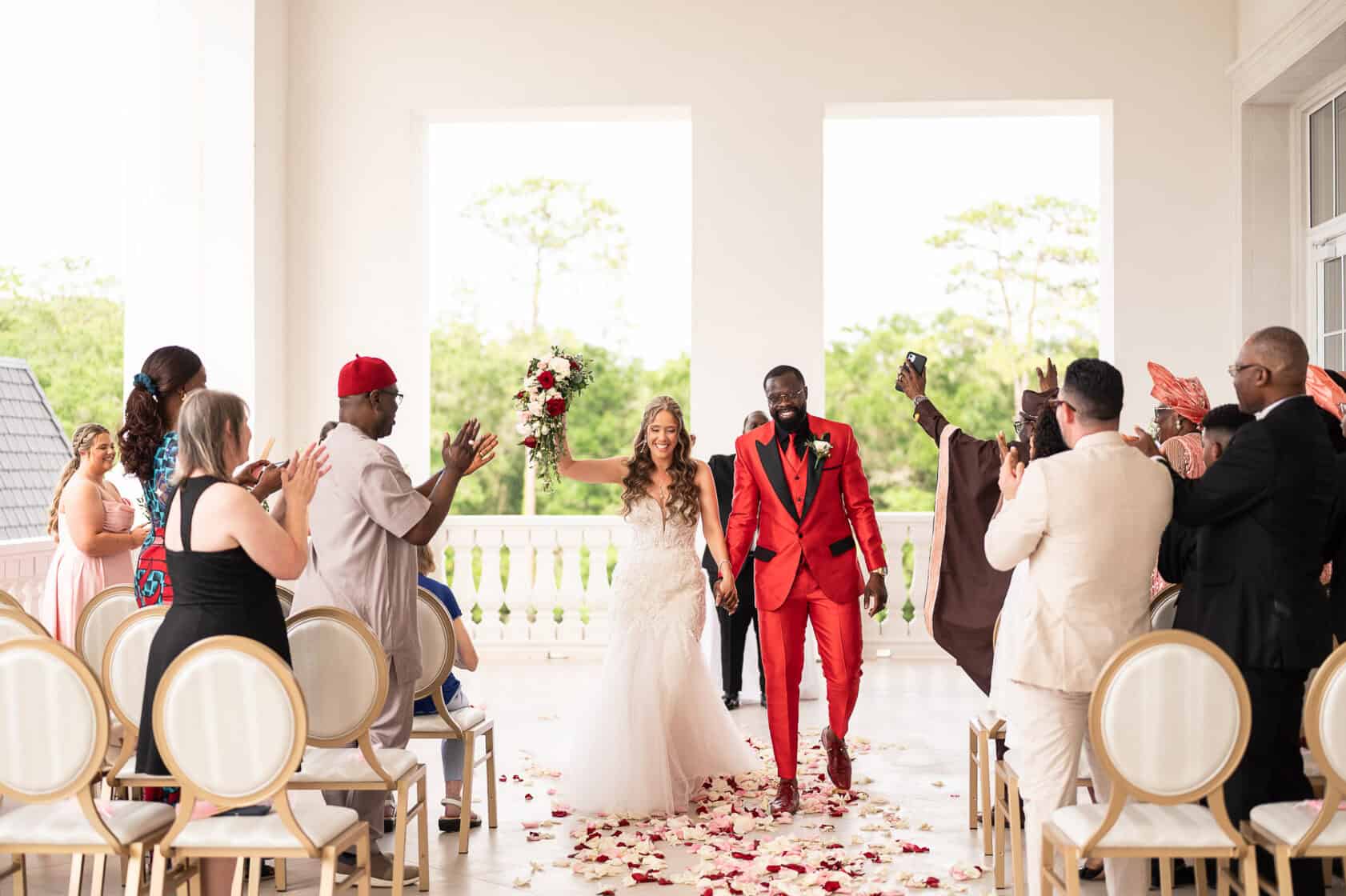 A bride in a white wedding gown and a groom in a red suit walk down an aisle covered in rose petals while wedding guests applaud.