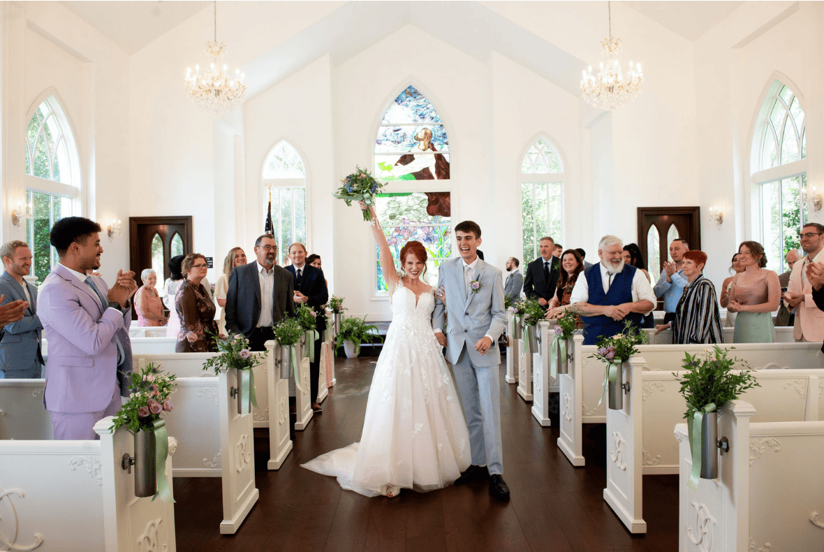 A bride and groom walk down the aisle of a chapel, the bride holding her bouquet in the air, with guests standing and clapping on both sides.