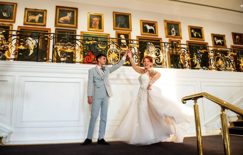 A bride and groom dancing on a mezzanine in front of a wall lined with framed portraits of dogs.