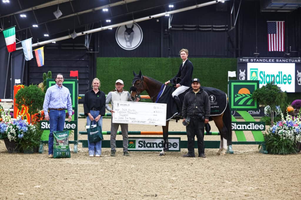 A group of four people and a horse posing in a show jumping arena with a large prize check.