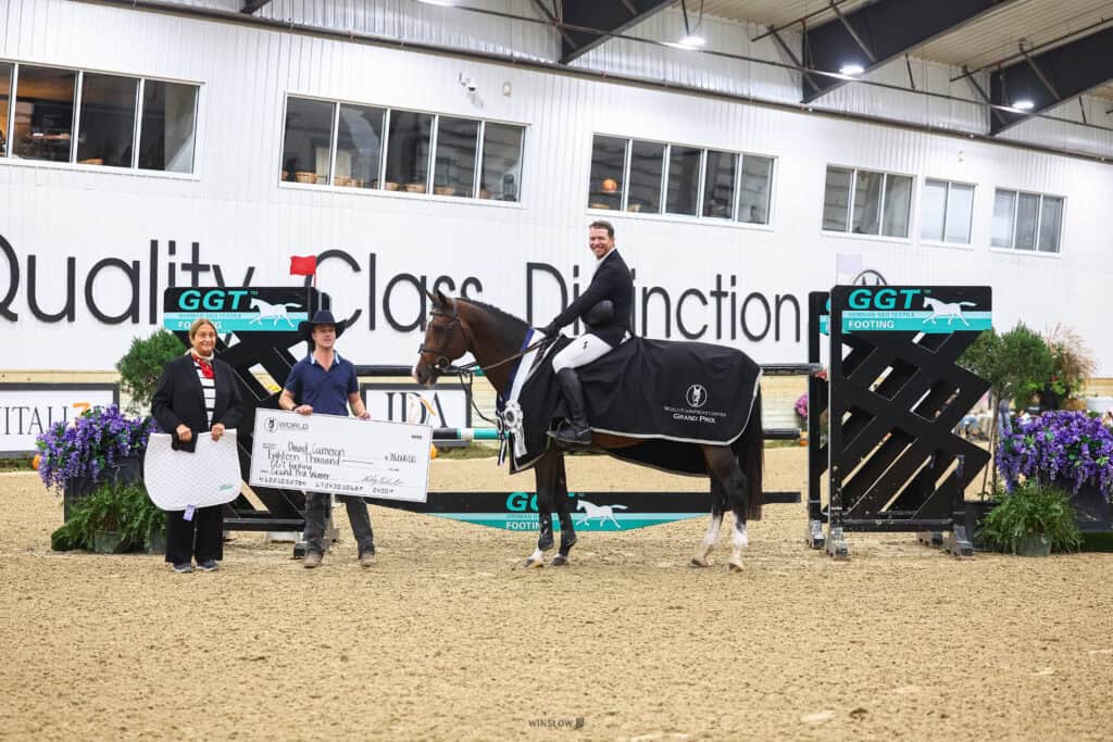 A man on a horse in an indoor arena stands next to a man and woman holding an oversized $18,000 check for a Grand Prix event.