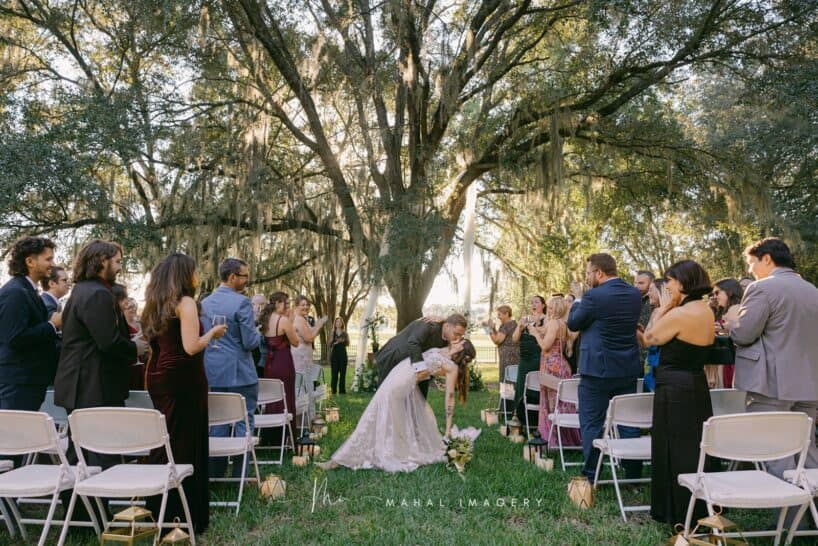 A bride and groom kiss as the groom dips her during their outdoor wedding ceremony, surrounded by guests in a grassy area under large trees.
