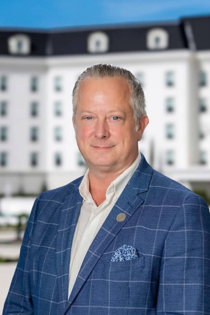 Headshot of a man wearing a blue plaid blazer, white shirt, and pocket square, with a building in the background.