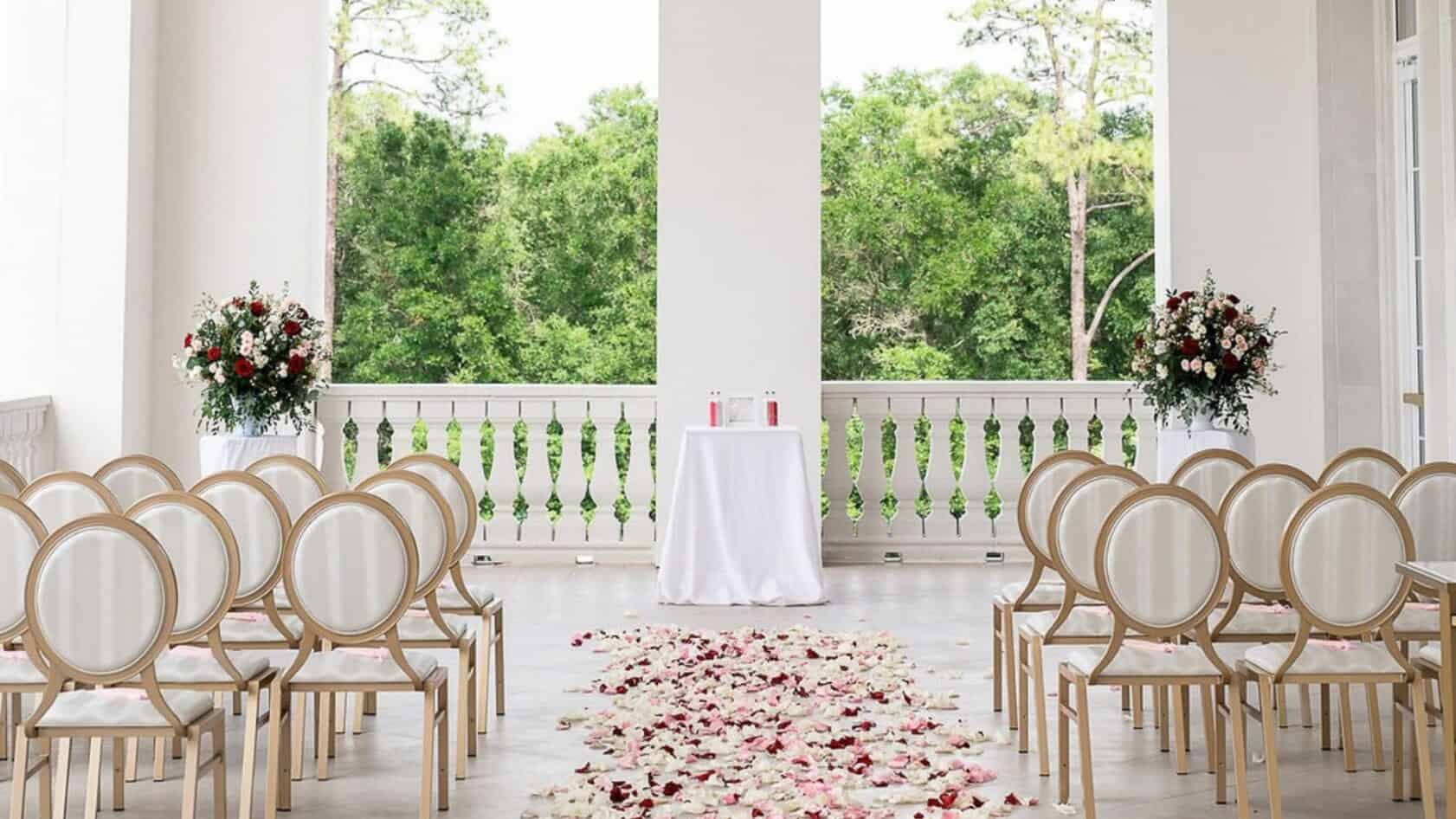 Rows of white chairs face an altar table and a path of flower petals on a balcony overlooking trees.