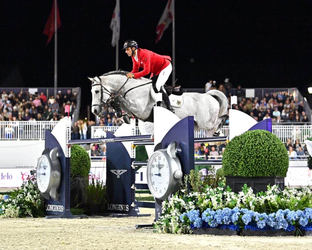 A show jumper in a red coat on a grey horse clearing a jump decorated with clock faces at an equestrian event.