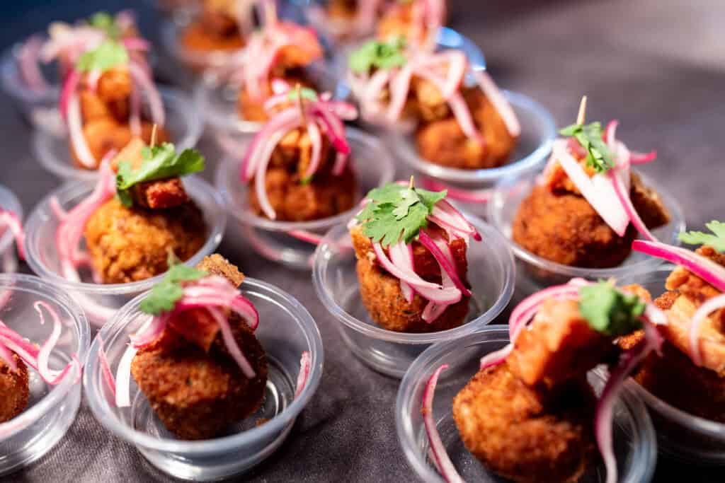 Small, breaded appetizers in clear plastic cups, topped with red onions and cilantro.