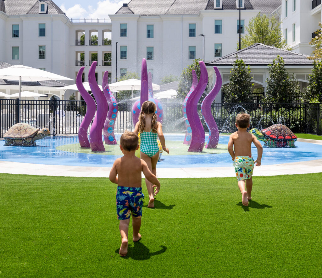 Three children run toward a splash pad featuring purple octopus tentacles and turtle statues.