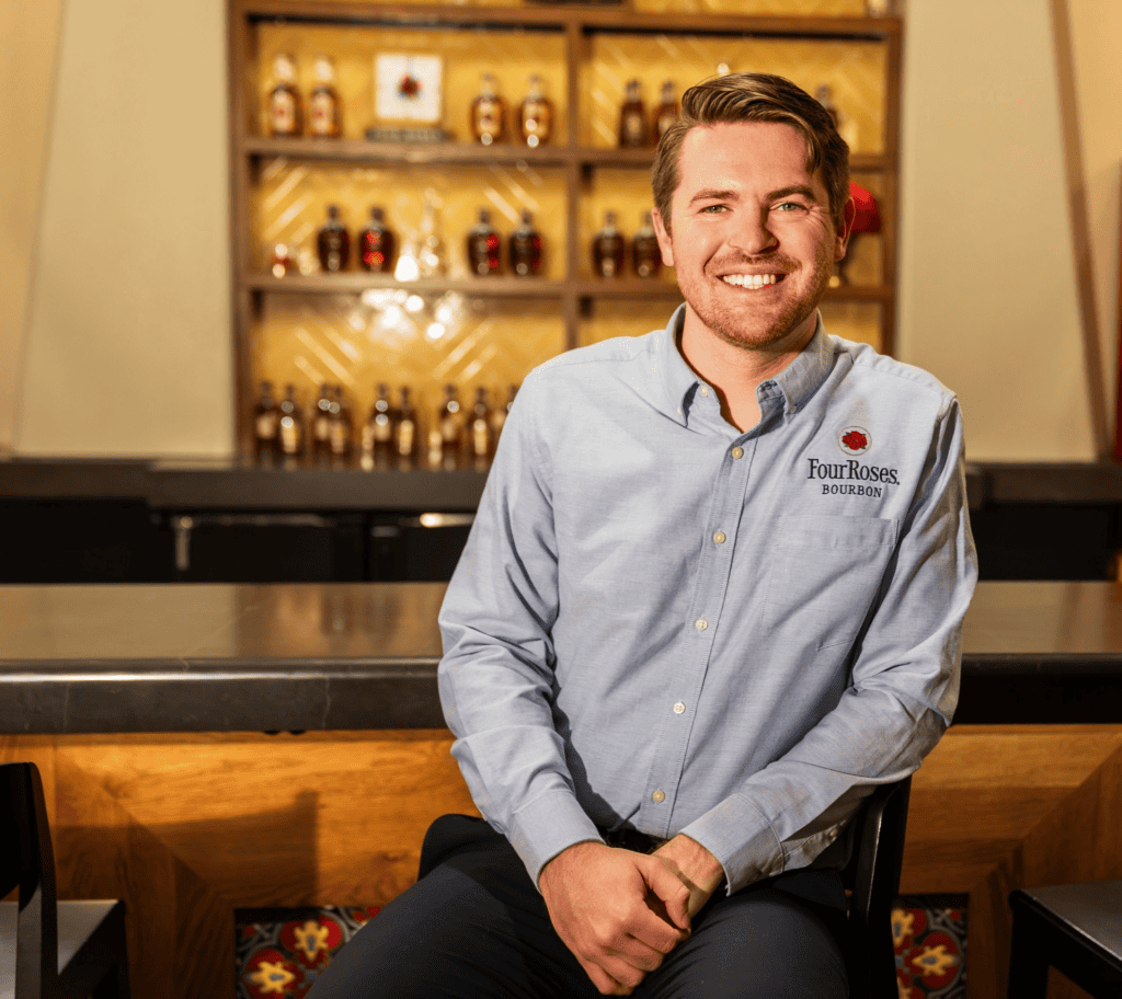 A smiling man in a light blue button-down shirt with the Four Roses Bourbon logo, sitting at a bar in front of shelves of liquor bottles.