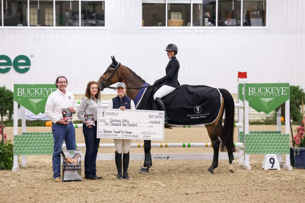 A horse and rider in equestrian gear stand with two people holding a large $10,500 check and animal feed bags at a Buckeye Nutrition sponsored show jumping arena.