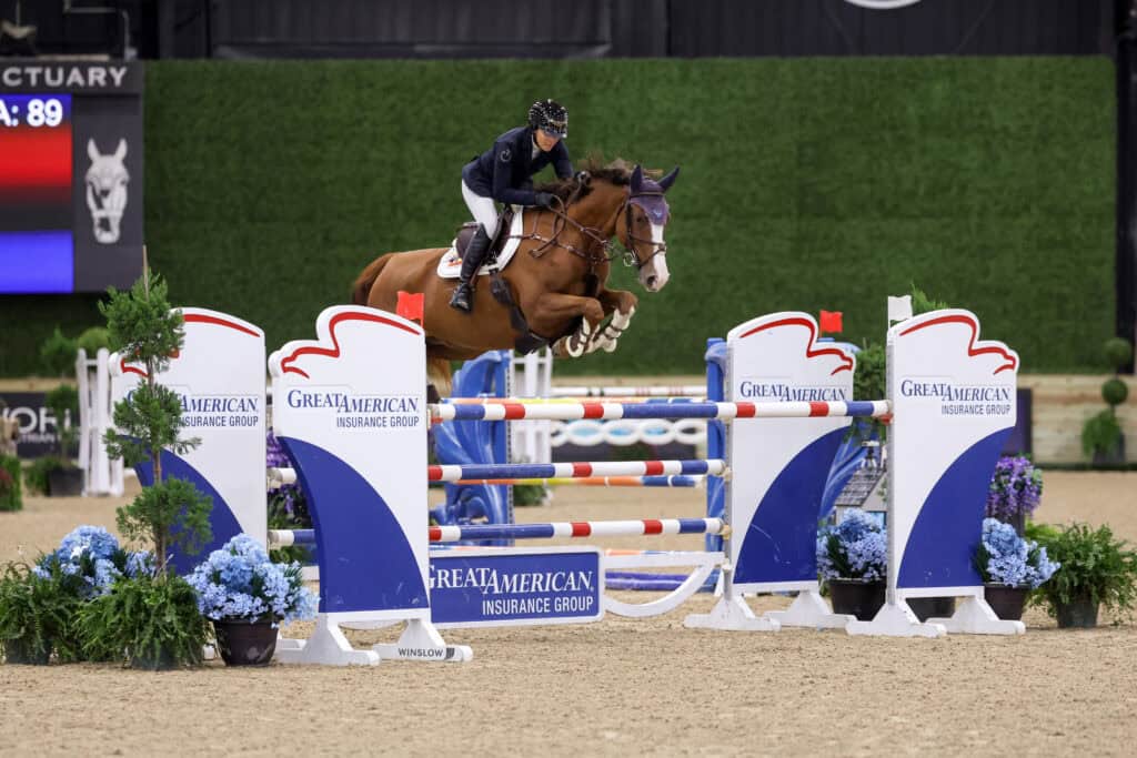 A horse and rider jumping over a blue, red, and white hurdle featuring Great American Insurance Group branding.
