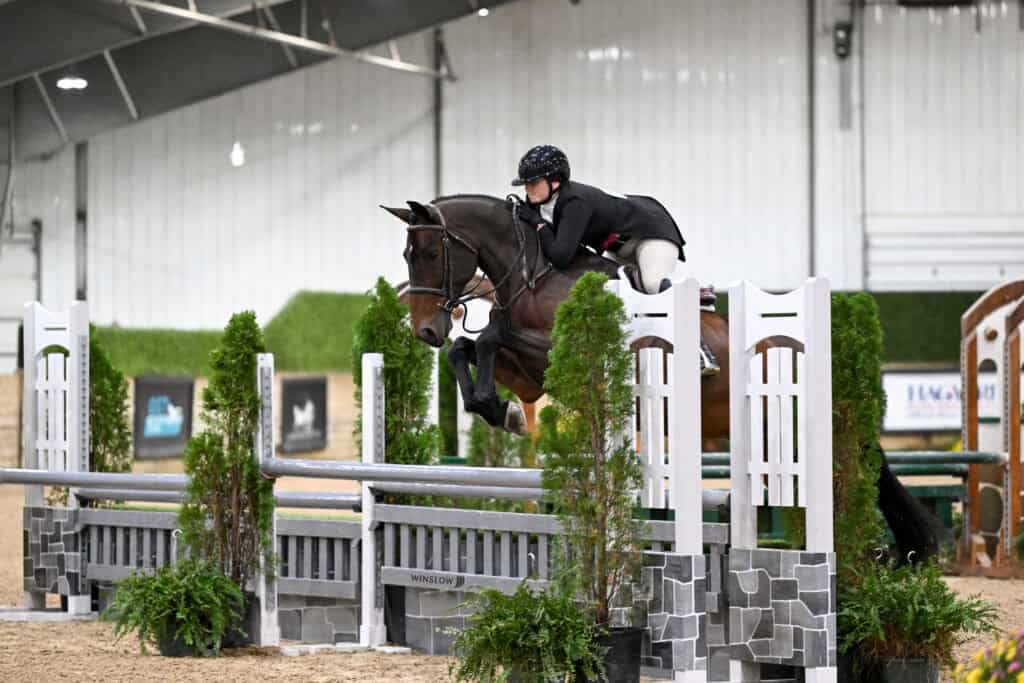 A rider in formal show attire jumps a horse over a white fence in an indoor arena.