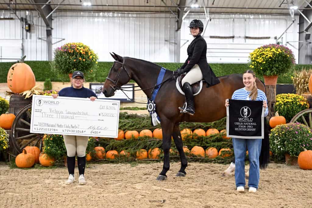 A rider on horseback stands between two women; one woman holds a $3,000 winning check, and the other holds a World Championship Hunter Derby banner.