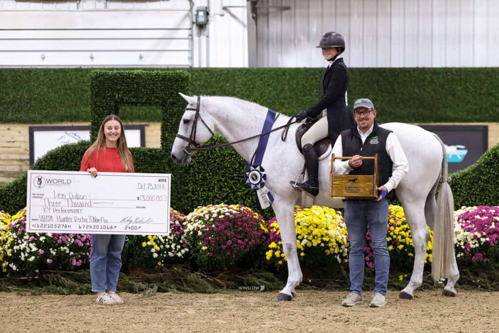 A woman in riding gear sits on a white horse, while a woman holds a large check and a man holds a wooden trophy box.