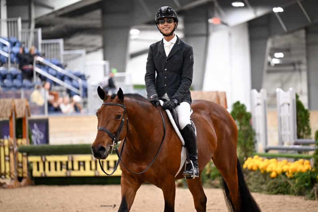 A smiling equestrian in riding gear mounted on a brown horse in an indoor arena.