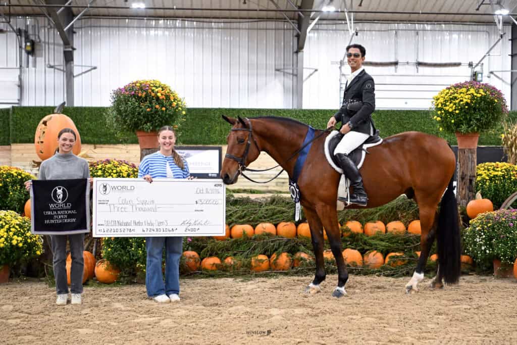 Two women stand in an indoor arena holding a $3,000 ceremonial check, while a man in equestrian attire sits on a horse wearing a blue ribbon.