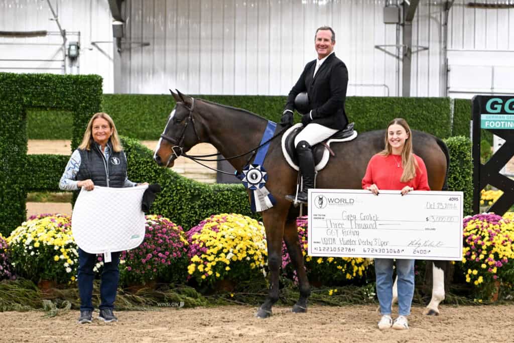 A rider on a brown horse wearing a blue ribbon stands between two people in an indoor arena, one holding a white saddle pad and the other holding a large check for three thousand dollars.
