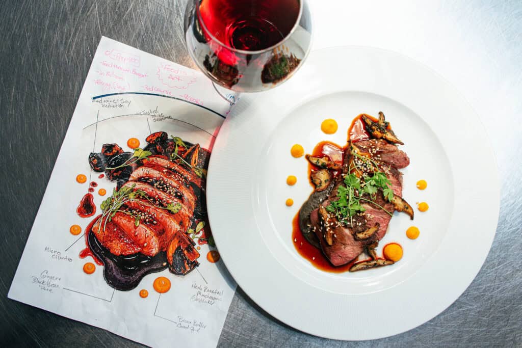 A plate of sliced steak with mushrooms and sauce next to a matching culinary sketch on paper and a glass of red wine.
