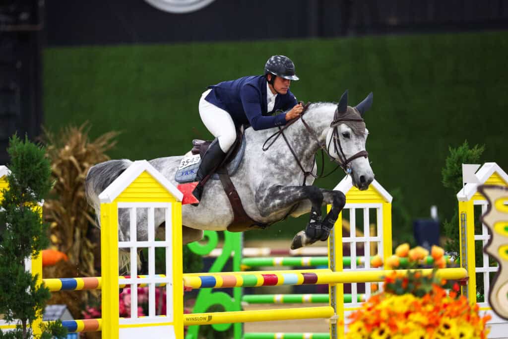 A show jumper on a grey horse clearing a yellow hurdle.